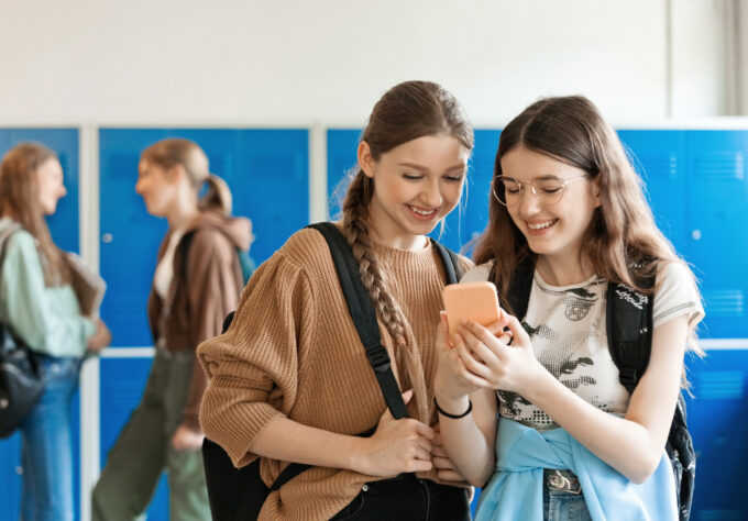 Two happy female high school students standing in school corridor against blue lockers and using mobile phone.