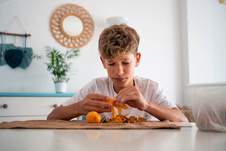 A teenager focuses on peeling fruits while seated at a sunny living room table. The apricots are fresh and vibrant. Surrounding him are bright decor and a mature potted plant.