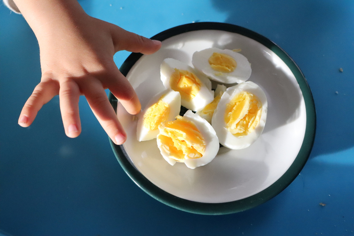 In the scene, a childs hand is seen reaching for boiled eggs in a bowl on a blue surface under bright sunlight