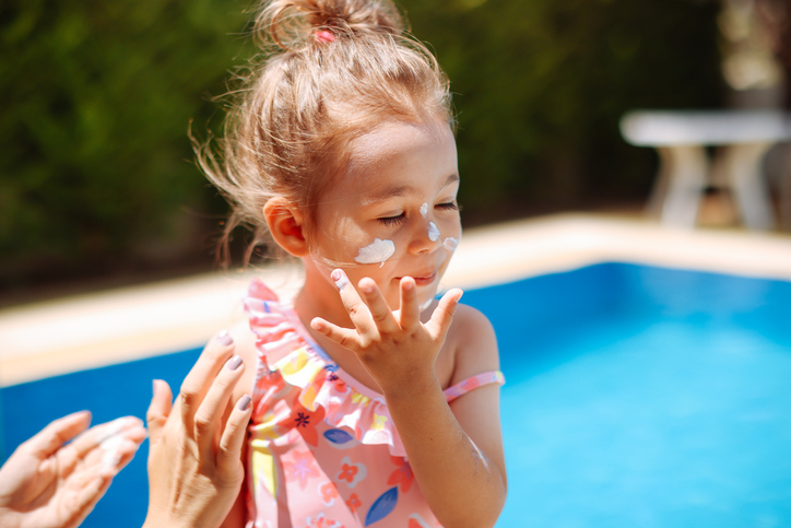 Mother taking care of her small kid while applying sunscreen to prevent sunburn. The kid is helping by applying sunscreen to her face.