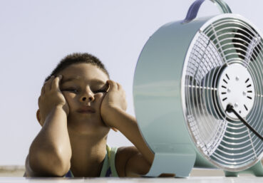 Overheated little child is sitting in front of electric fan trying to cool down.