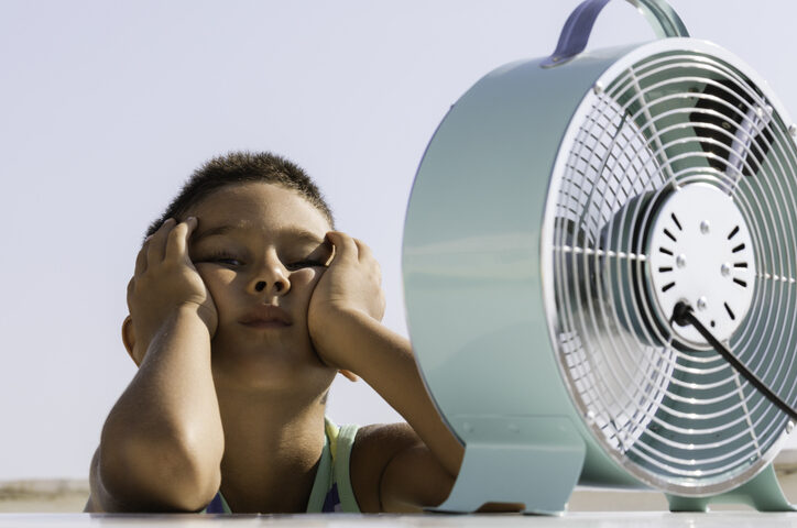 Overheated little child is sitting in front of electric fan trying to cool down.