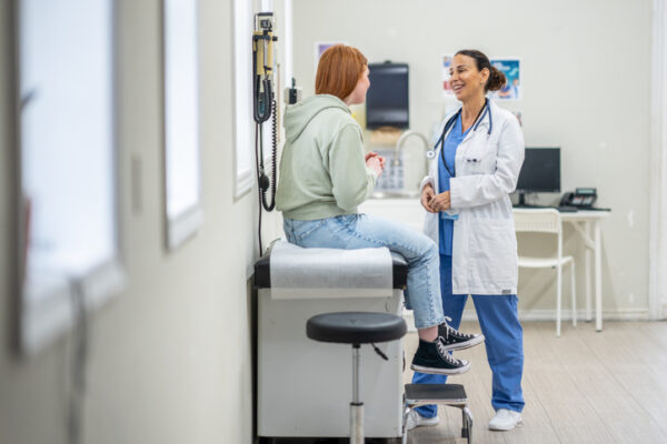 A young female teenager sits up on an exam table during a routine medical appointment with her doctor. She is dressed casually and talking with her doctor who is standing in front of her as they talk.