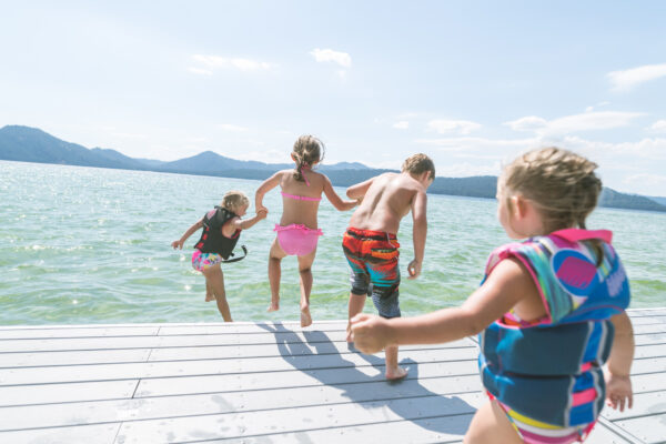 Four kids jumping into a lake on a summer day with two of the younger children wearing life vests to demonstrate proper water safety