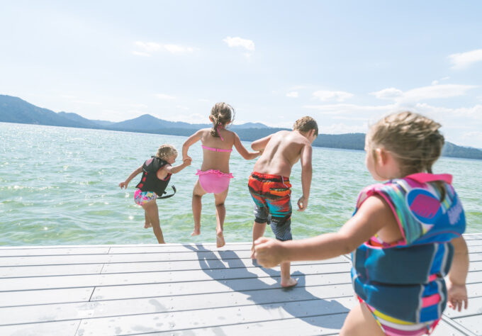 Four kids jumping into a lake on a summer day with two of the younger children wearing life vests to demonstrate proper water safety