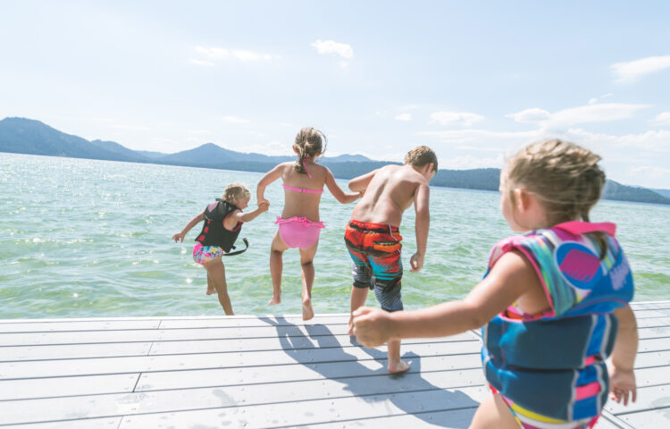 Four kids jumping into a lake on a summer day with two of the younger children wearing life vests to demonstrate proper water safety