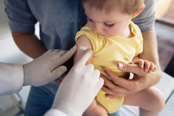 Unrecognizable doctor putting a patch on the little boy's shoulder after successful RSV vaccination. Little boy sitting in father´s lap and looking at the patch.