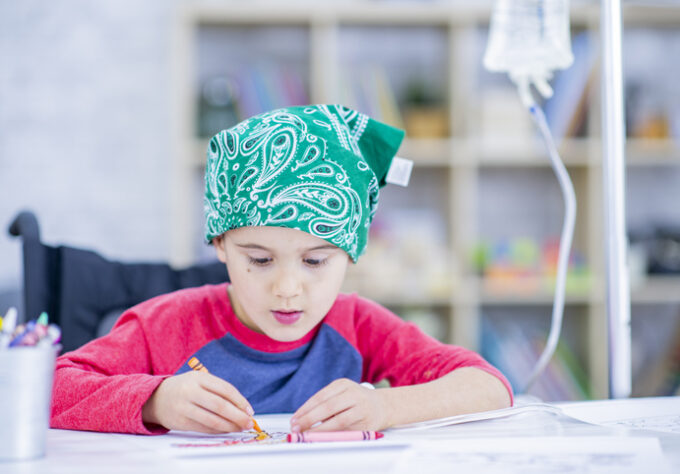 A young boy with a bandana and an IV drip beside him draws with crayons. He in indoors, sitting at a desk in a treatment centre.