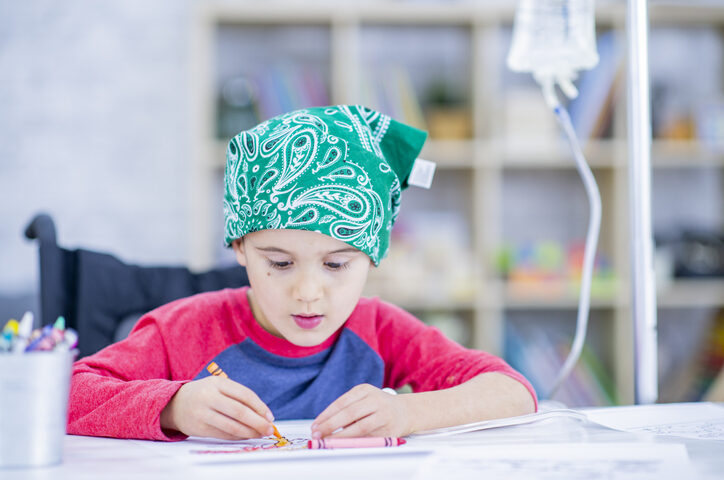 A young boy with a bandana and an IV drip beside him draws with crayons. He in indoors, sitting at a desk in a treatment centre.