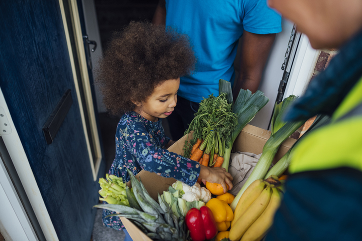 Over the shoulder view of a person delivering a box full of fresh vegetables to a family who has a child with cancer.