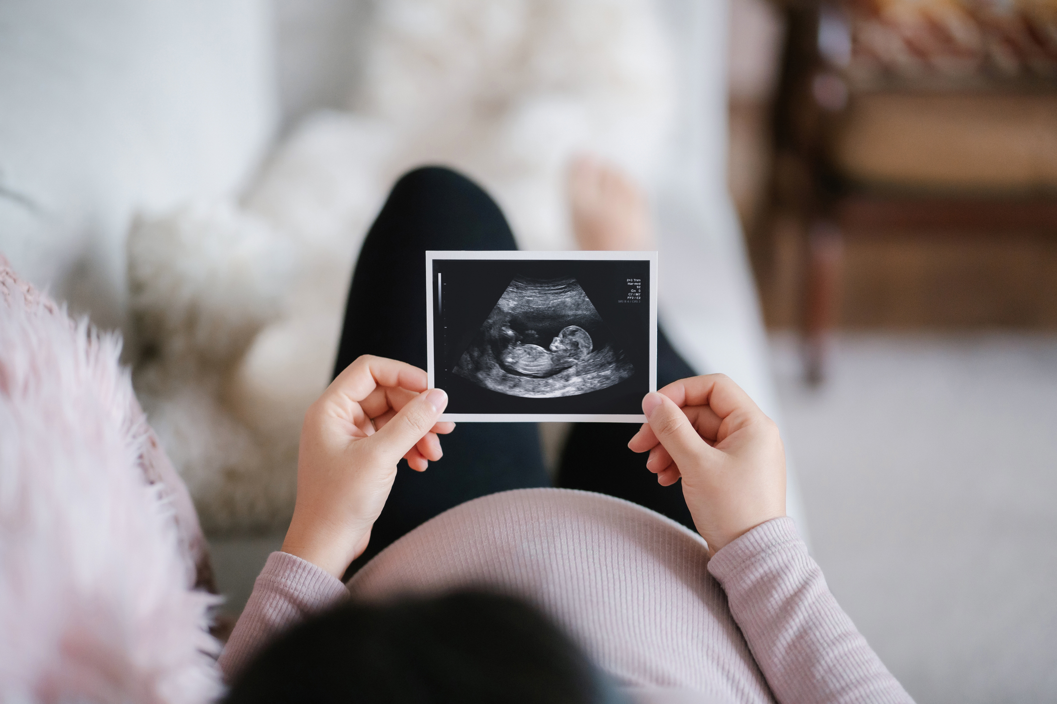 Young pregnant woman lying on sofa at home, looking at the ultrasound scan photo of her baby.