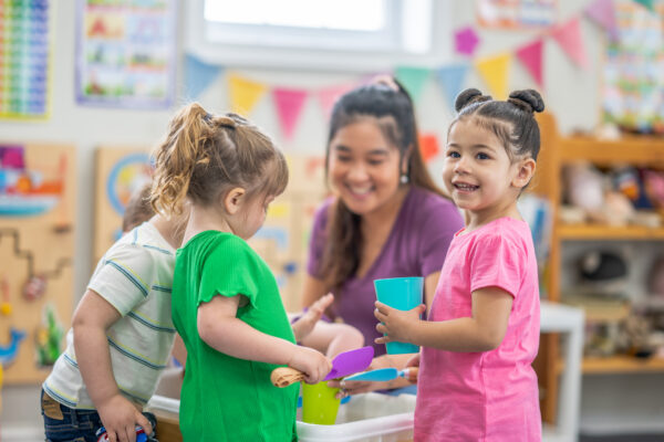 A preschool teacher knees down with a small group of children as they gather around an activity table filled with rice. They are each dressed casually and are smiling as they enjoy playing together.