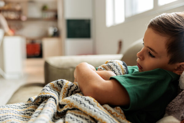 Young boy lying on the couch covered in a blanket, looking sad while recovering from illness in the living room.