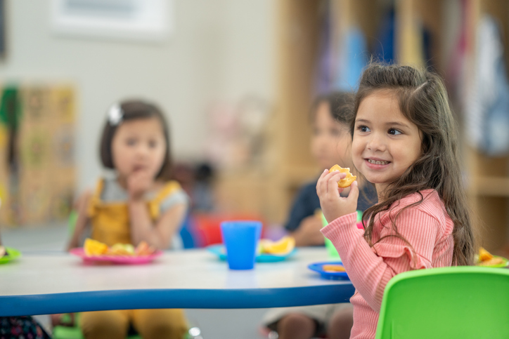 A small group of preschool students sit at a table as they enjoy a snack together.