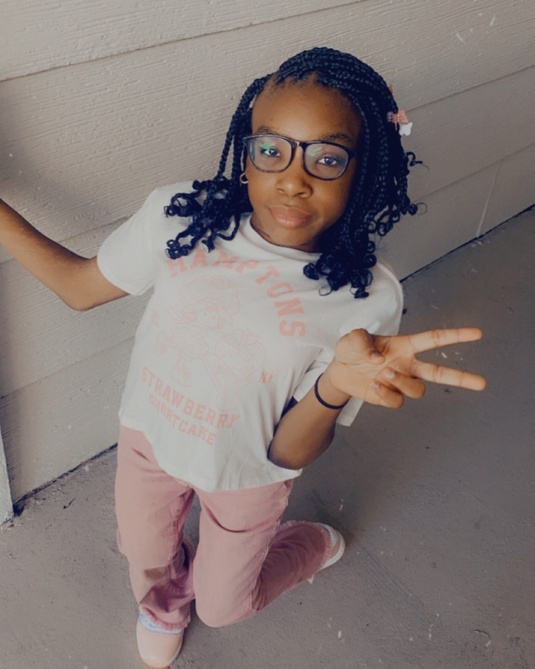 Girl with glasses and a white shirt posing and holding up a peace sign in front of a white wall.