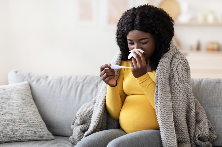 Pregnant woman wrapped in blanket feeling sick with high fever, sitting on couch at home and measuring temperature with thermometer to decide whether or not to call her doctor.