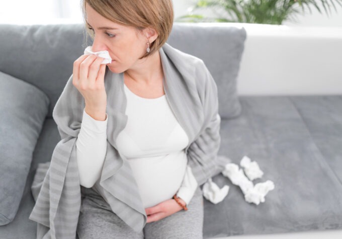 Pregnant woman catching cold, flu, virus, sitting at home on a couch, blowing nose and being sick.