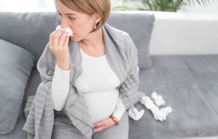 Pregnant woman catching cold, flu, virus, sitting at home on a couch, blowing nose and being sick.