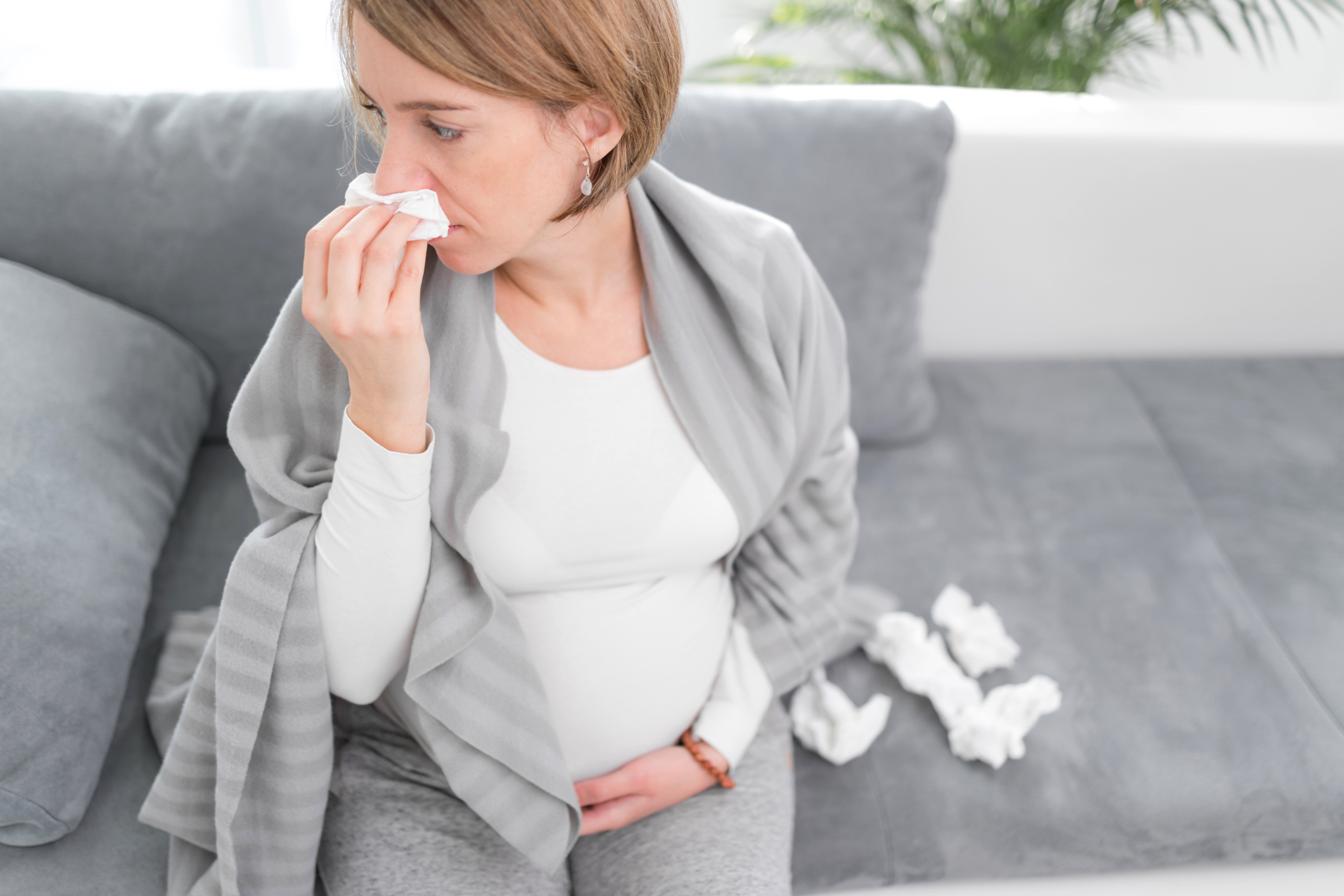 Pregnant woman catching cold, flu, virus, sitting at home on a couch, blowing nose and being sick.