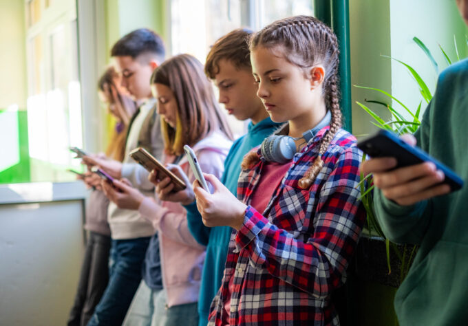 Teenage students are standing in the school hallway, all looking at news and social media on their phones.