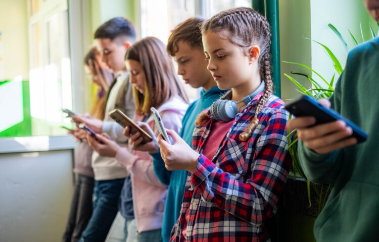 Teenage students are standing in the school hallway, all looking at news and social media on their phones.