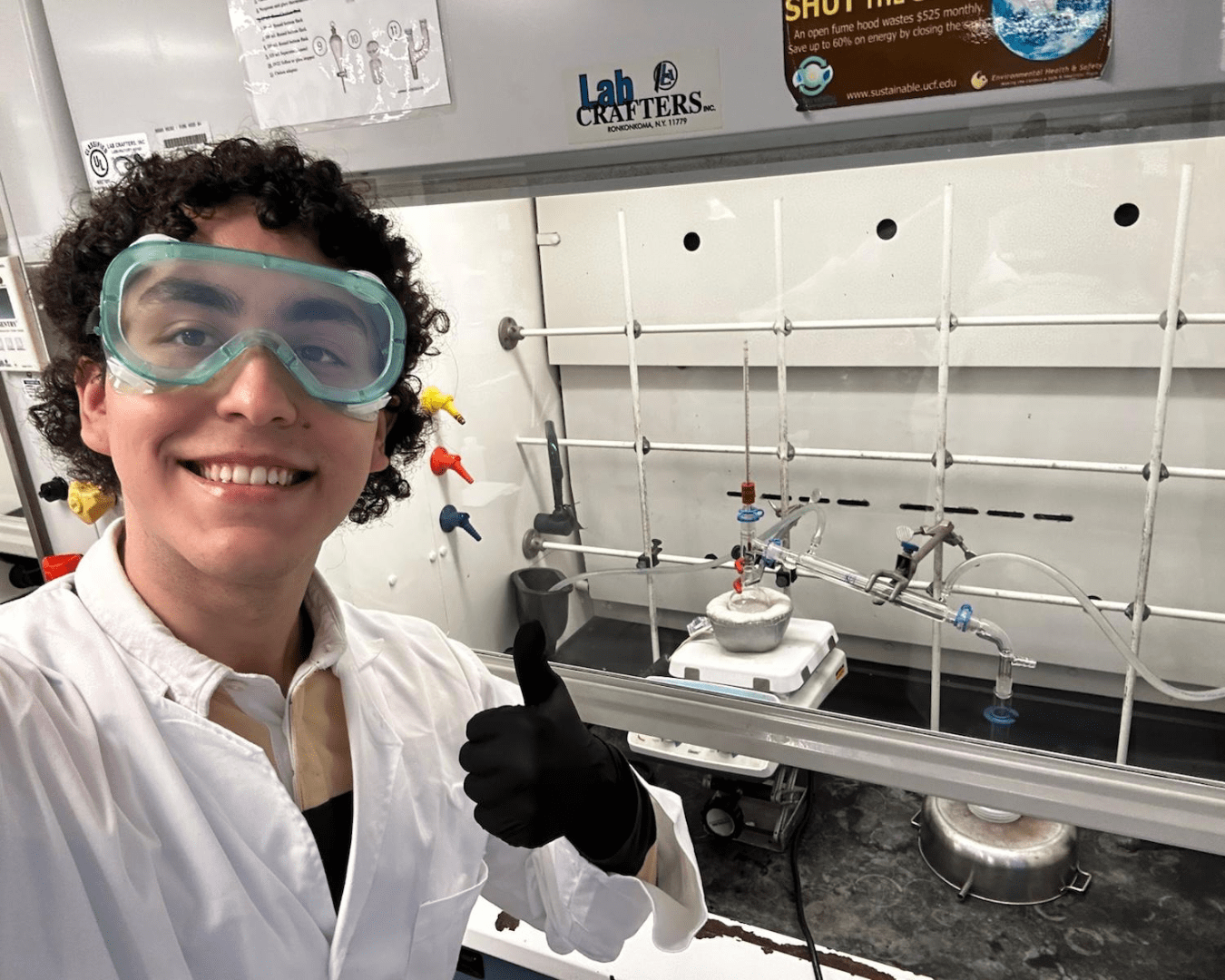 Teen male in goggles and medical jacket smiles for a selfie while in an educational lab