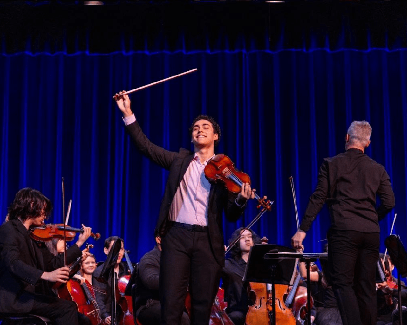 Teen male smiling and standing on stage in front of an orchestra after finishing a violin solo