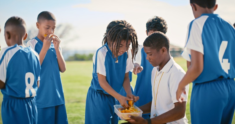 Snack break, soccer and children with a coach at sports practice, game or match. Fruit diet, eating and young football players with food for nutrition during a competition or training with a man