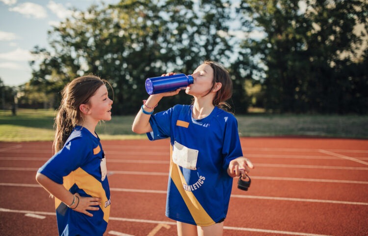 Two little girls are having a water break on the running track at athletics club. One girl is standing with her hands on hips, the other is taking a big gulp of water from bottle.