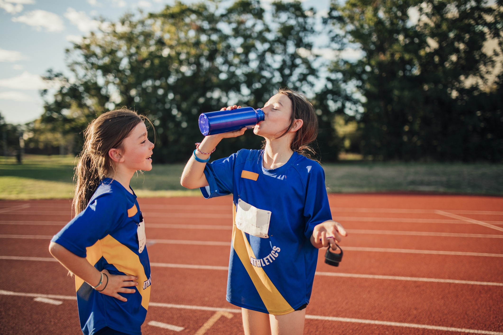 Two little girls are having a water break on the running track at athletics club. One girl is standing with her hands on hips, the other is taking a big gulp of water from bottle.