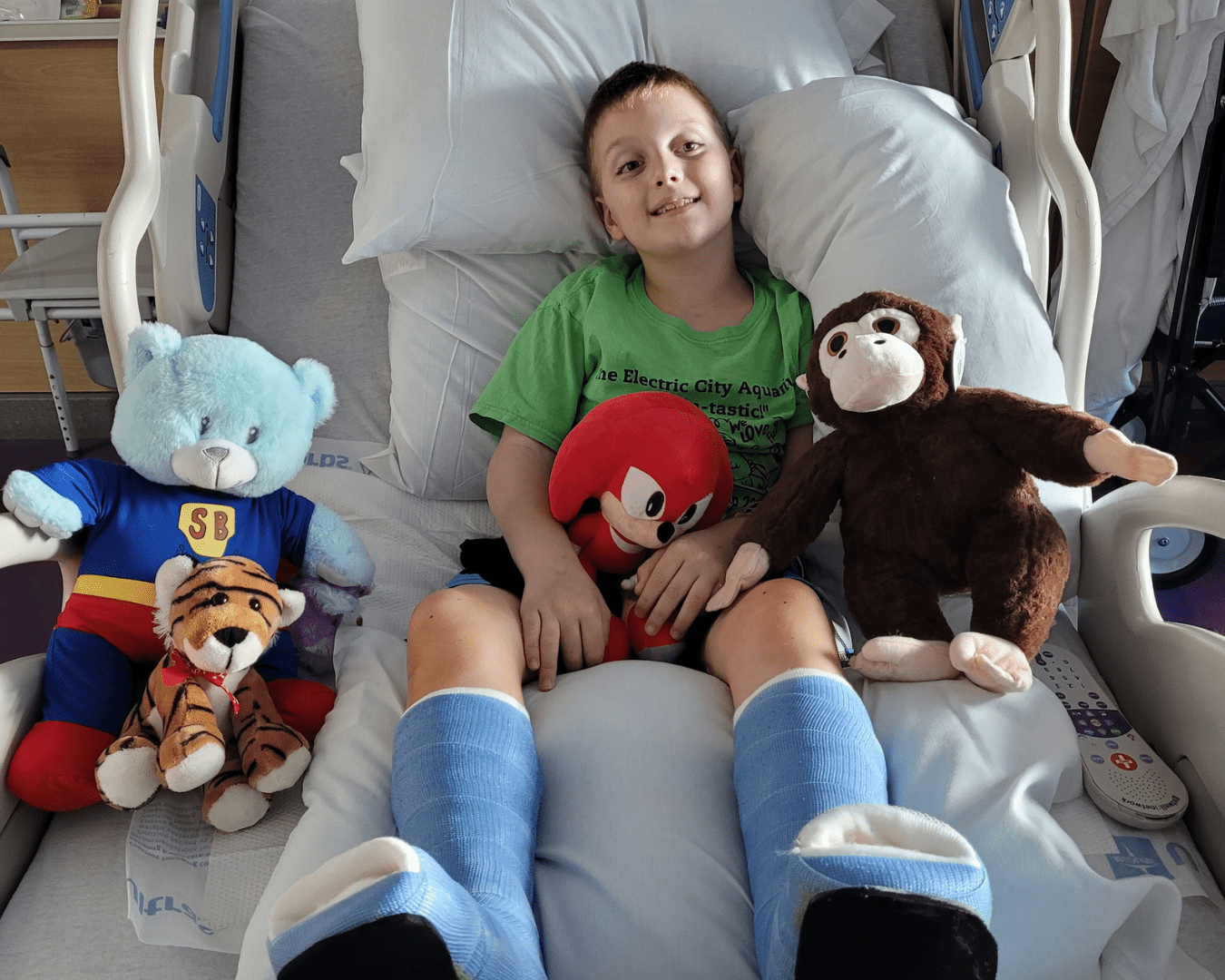 Boy with casts on both legs smiles for a photo while in a hospital bed surrounded by stuffed animals.