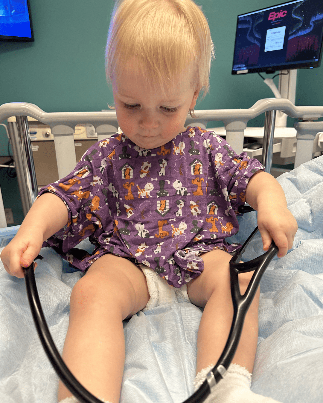 Young blonde child in purple hospital gown plays with a stethoscope while sitting in a hospital bed.