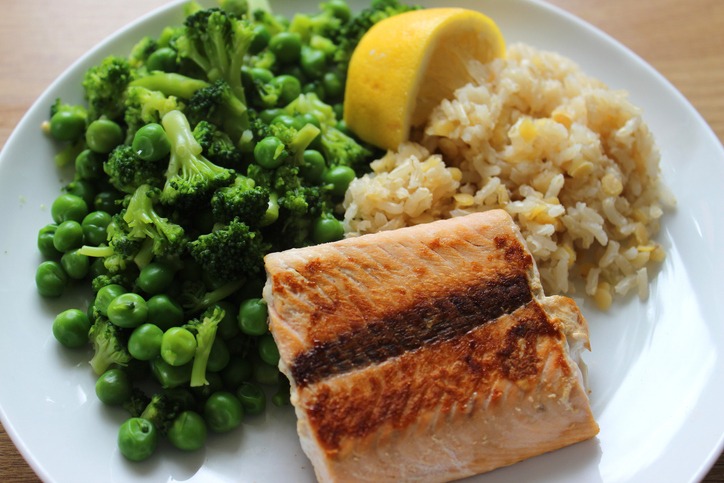 Photo showing a healthy meal consisting of a dry fried salmon fillet served with brown rice and red lentils, garden peas / frozen peas mixed with small pieces of broccoli and a slice of lemon.