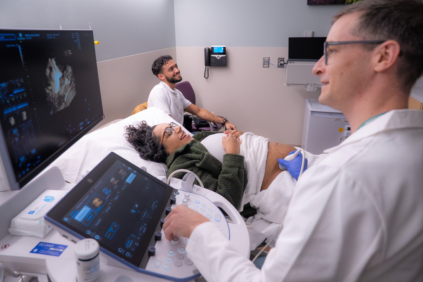 Image of women lying down with a smile on her face with her partner close by while getting a fetal ultrasound and the doctor examines the imaging on a screen.
