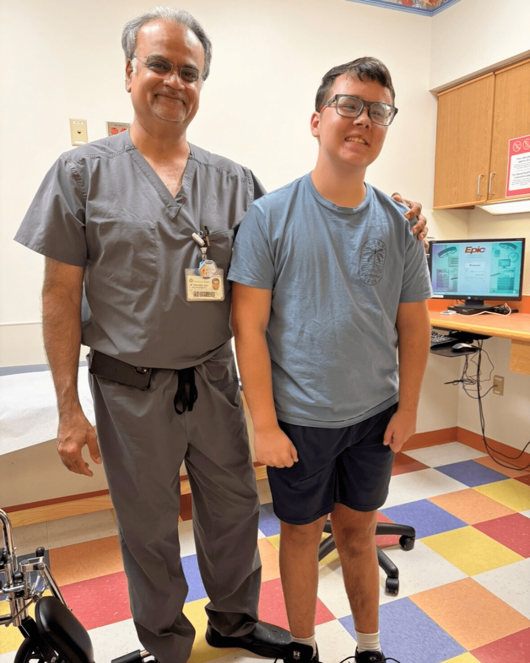 Boy with glasses in a blue T-shirt and shorts smiles for a photo with his doctor. Doctor is wearing grey scrubs and they are standing in a clinic room.