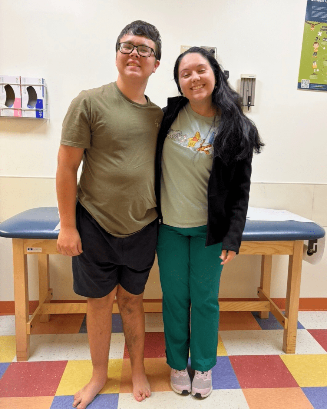 Teen boy with classes smiles for a photo with a clinican who is wearing green scrubs. Photo was taken in a clinic room.