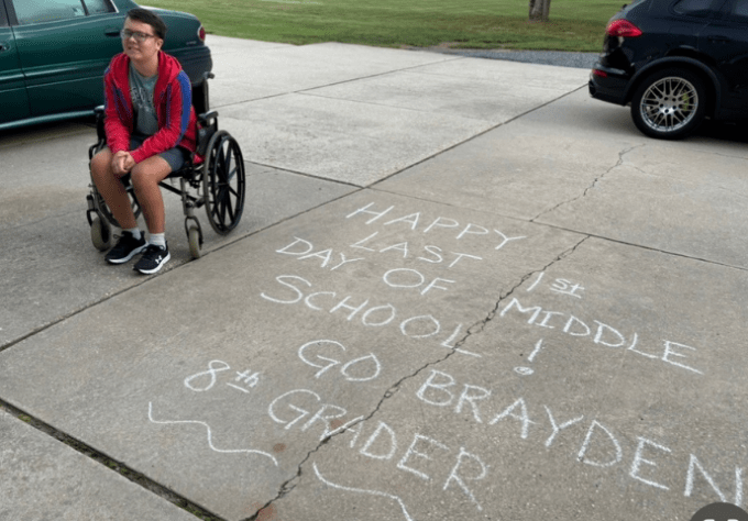 Boy wearing a red jacket and sitting in a wheelchair smiles for a photo in a driveway next to a chalk writing that reads, "Happy Last 1st Day of Middle School! Go Brayden!"