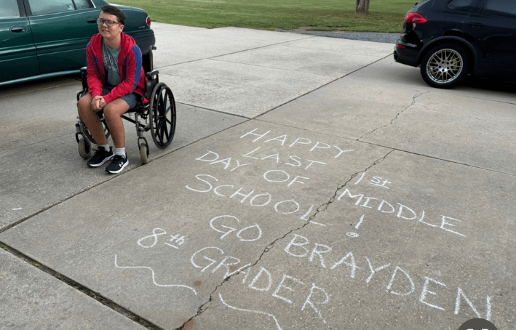 Boy wearing a red jacket and sitting in a wheelchair smiles for a photo in a driveway next to a chalk writing that reads, "Happy Last 1st Day of Middle School! Go Brayden!"