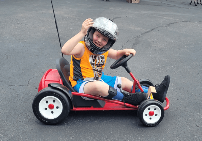 Boy with helmet on smiles and waves for a photo while riding in a go kart