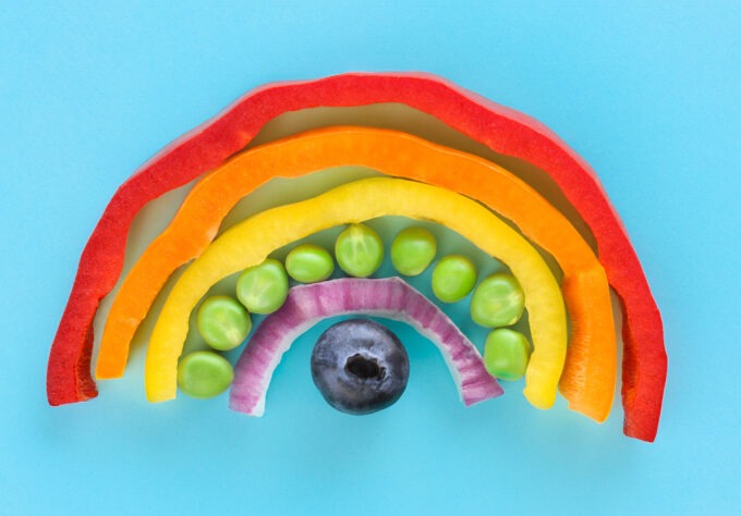Photo showing close-up, elevated view of arch of chopped multi-coloured fruits and vegetables. Rainbow of chopped red, orange and yellow bell peppers, green peas, red onion and a blueberry.
