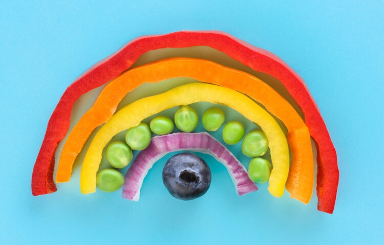 Photo showing close-up, elevated view of arch of chopped multi-coloured fruits and vegetables. Rainbow of chopped red, orange and yellow bell peppers, green peas, red onion and a blueberry.