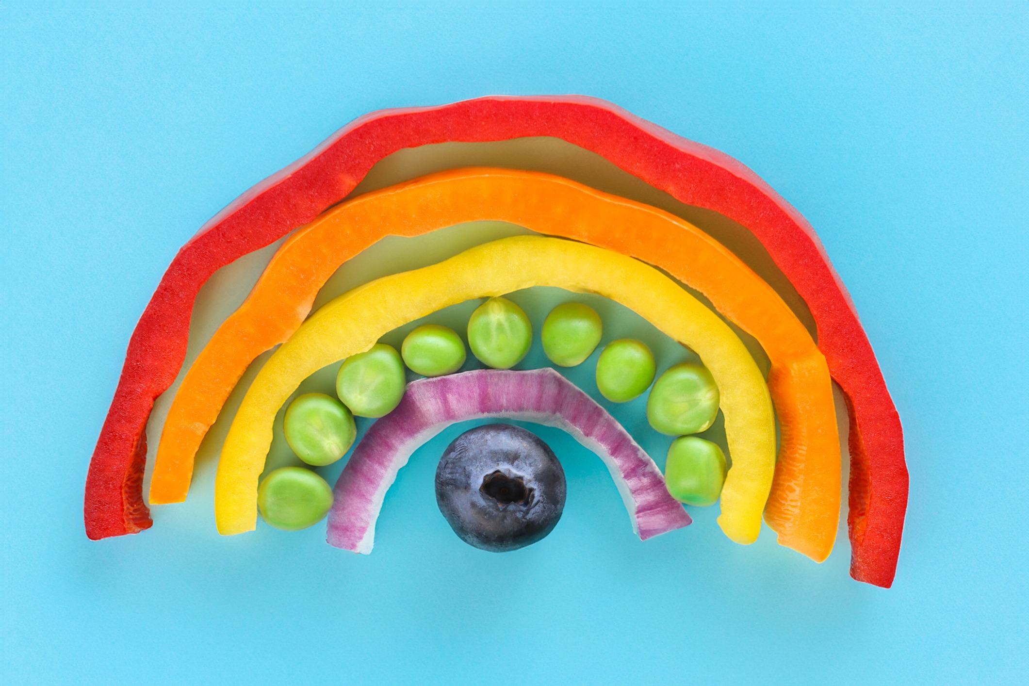 Photo showing close-up, elevated view of arch of chopped multi-coloured fruits and vegetables. Rainbow of chopped red, orange and yellow bell peppers, green peas, red onion and a blueberry.