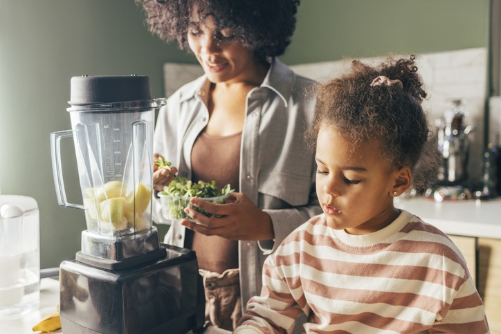 A mother and her daughter preparing a healthy juice made of fruits and vegetables using a blender.
