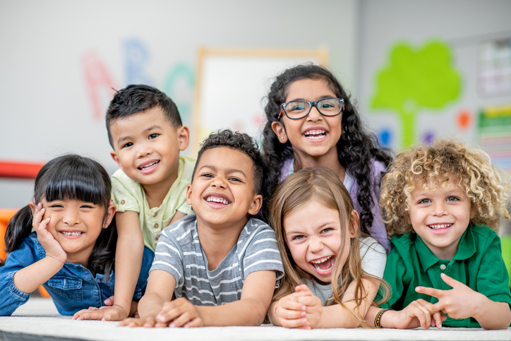 A small group of children lay tightly together on the floor of their classroom as they pose for a portrait. They are each dressed comfortably and are smiling.