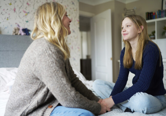 Mother and daughter sit together on a bed holding hands as they have a supportive conversation.