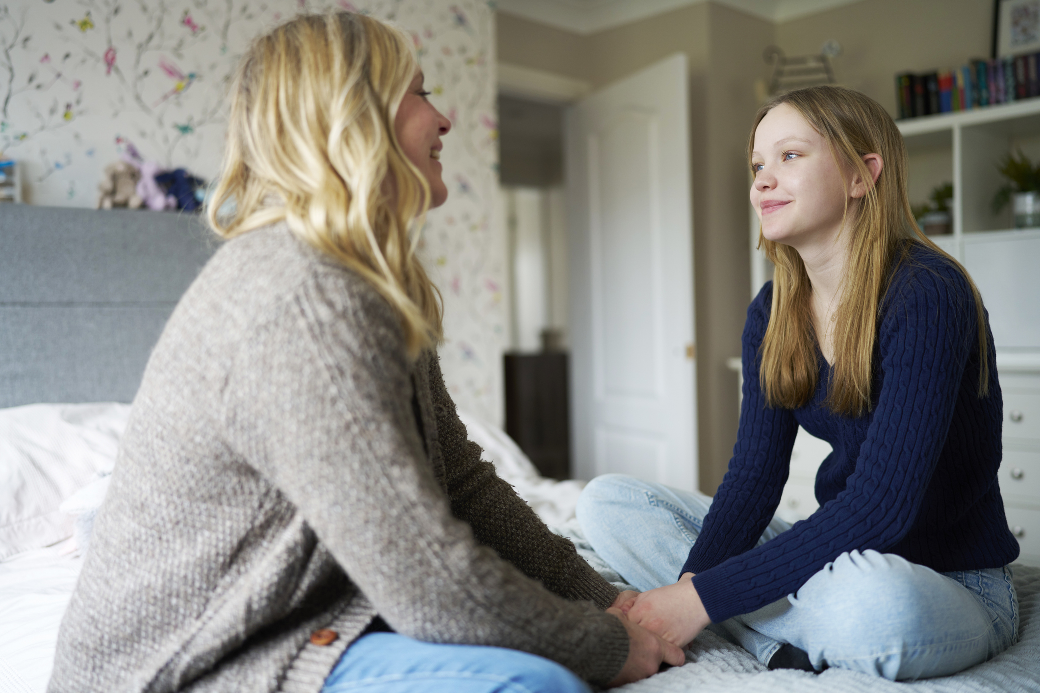 Mother and daughter sit together on a bed holding hands as they have a supportive conversation.