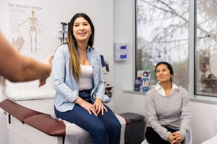 Teen girl sits in doctor's office on an exam table discussing her PMOS symptoms with doctor with the support of her mother who is sitting in on the visit.