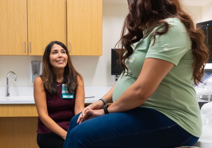 Pregnant woman sits in doctors office while a maternal fetal medicine specialist listens to her questions.