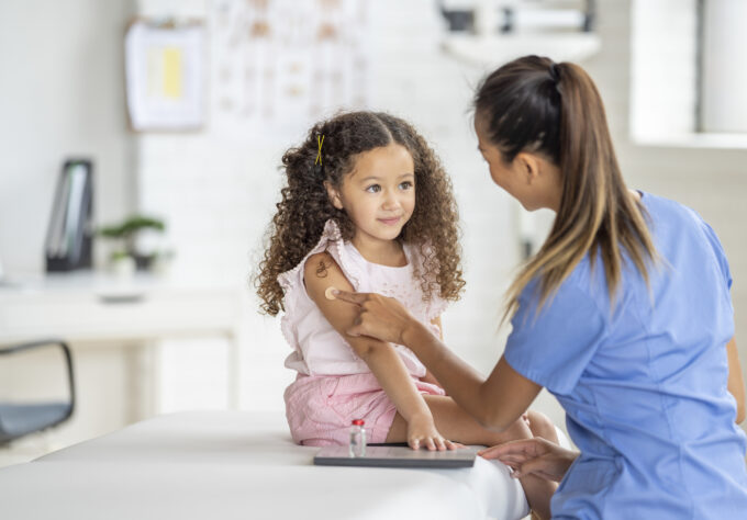 Healthcare professional placing a bandage on a young child’s upper arm after an immunization while the child sits on an examination table in a medical office.