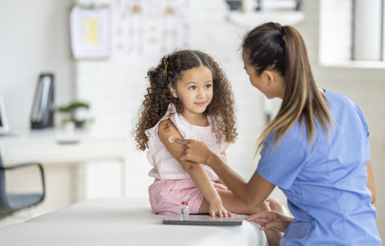 Healthcare professional placing a bandage on a young child’s upper arm after an immunization while the child sits on an examination table in a medical office.